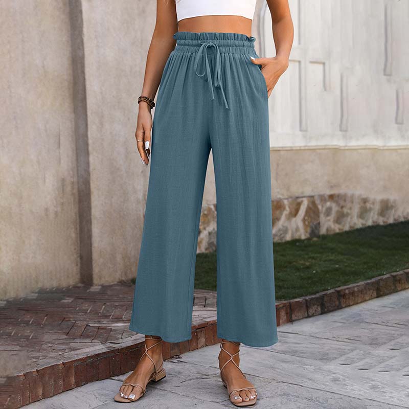 Frau in blauer, weiter Leinenhose und weißem Crop-Top, steht auf Gehweg. Sommermode, lässiger Stil, bequeme Kleidung, modische Sandalen.
