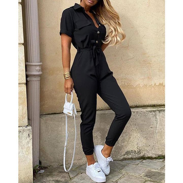 Frau in schwarzem Jumpsuit mit weißen Sneakers und kleiner weißer Handtasche, lehnt an einer Wand. Modetrend, lässiger Stil, Streetwear.