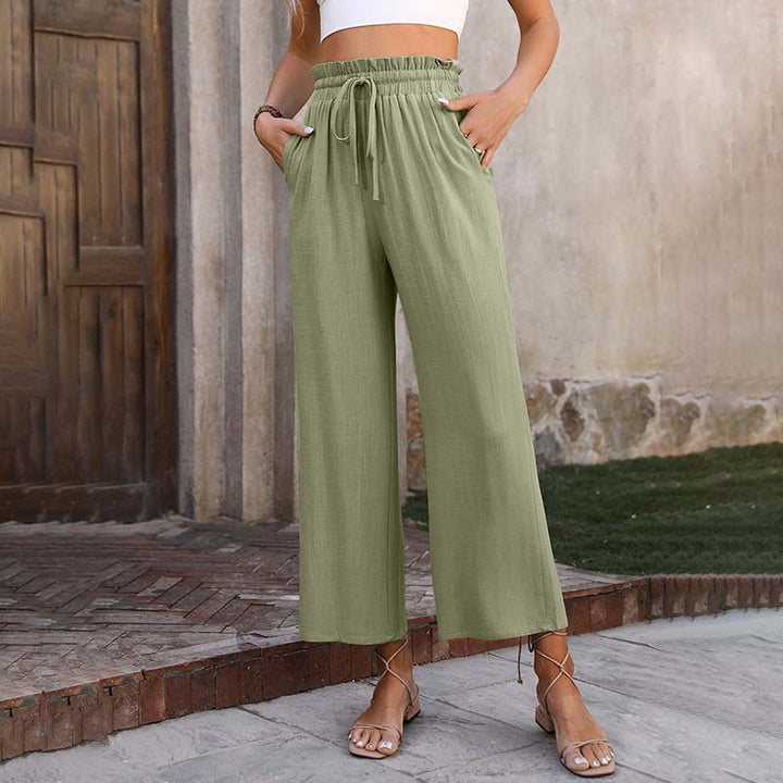 Frau trägt grüne Leinenhose mit weitem Bein und Kordelzug, steht vor rustikaler Wand. Modetrend, Sommeroutfit, Damenbekleidung.
