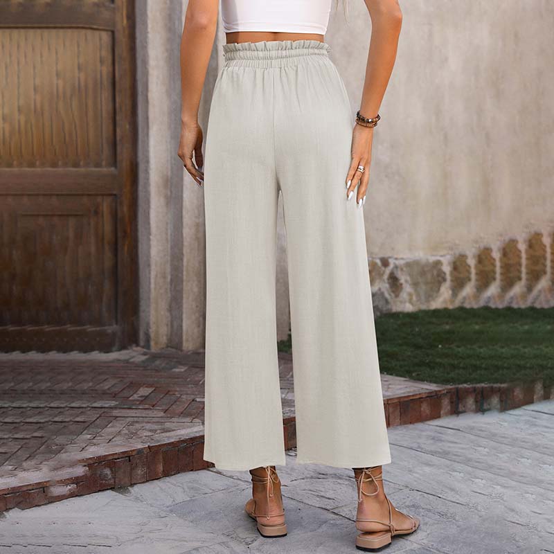 Frau in beiger, weiter Leinenhose und weißem Top, steht vor Holztür. Sommermode, lässiger Stil, Sandalen. Ideal für warme Tage.