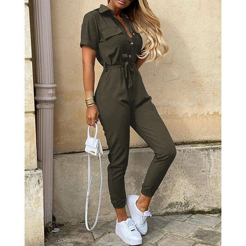 Frau in olivgrünem Jumpsuit mit weißen Sneakers und kleiner weißer Handtasche, steht vor beiger Wand. Modetrend, lässiger Stil, Streetwear.