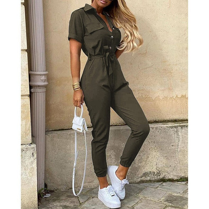 Frau in olivgrünem Jumpsuit mit weißen Sneakers und kleiner weißer Handtasche, steht vor beiger Wand. Modetrend, lässiger Stil, Streetwear.