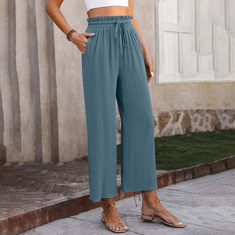 Frau in blauer, locker sitzender Hose und Sandalen vor einer Steinmauer. Modetrend, Sommeroutfit, Damenbekleidung, lässiger Stil.