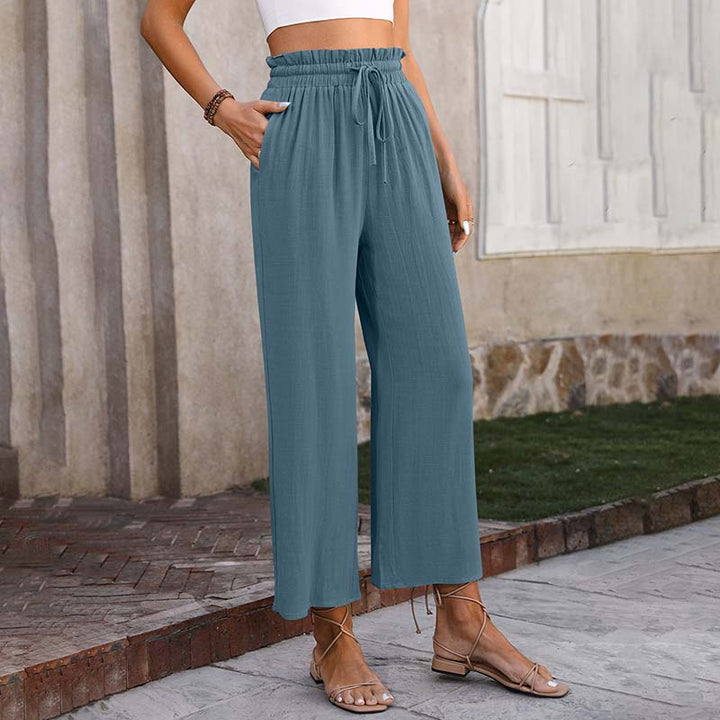 Frau in blauer, locker sitzender Hose und Sandalen vor einer Steinmauer. Modetrend, Sommeroutfit, Damenbekleidung, lässiger Stil.