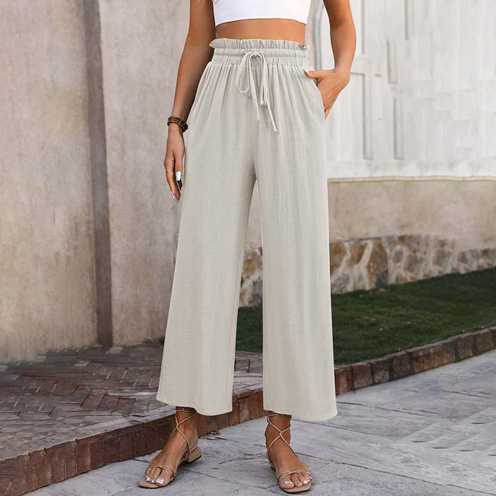 Frau trägt beige, weite Leinenhose mit hoher Taille und Kordelzug, kombiniert mit weißem Crop-Top und Sandalen. Sommermode, lässiger Stil.