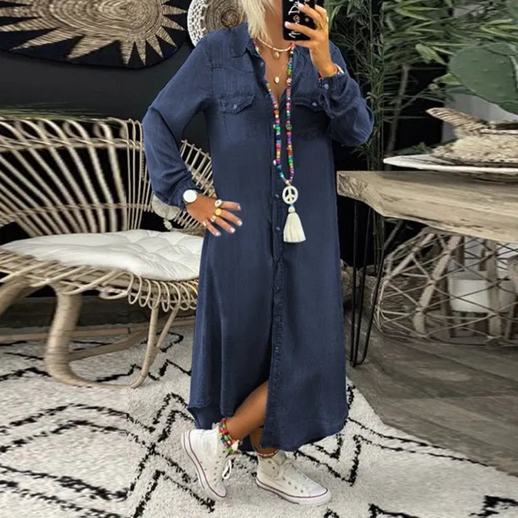 Frau in langem, blauem Jeanskleid mit buntem Schmuck und weißen Sneakers in modernem Wohnzimmer mit Rattanmöbeln und Pflanzen.