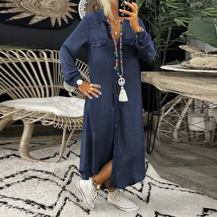 Frau in langem, blauem Jeanskleid mit buntem Schmuck und weißen Sneakers in modernem Wohnzimmer mit Rattanmöbeln und Pflanzen.