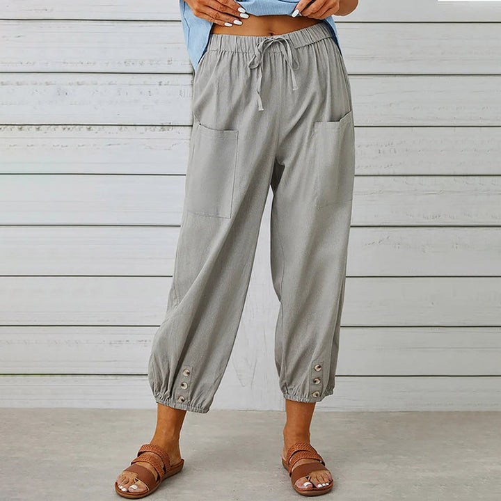 Damenhose aus Leinen in Grau mit elastischem Bund und Knöpfen an den Knöcheln, kombiniert mit braunen Sandalen vor weißer Holzwand.