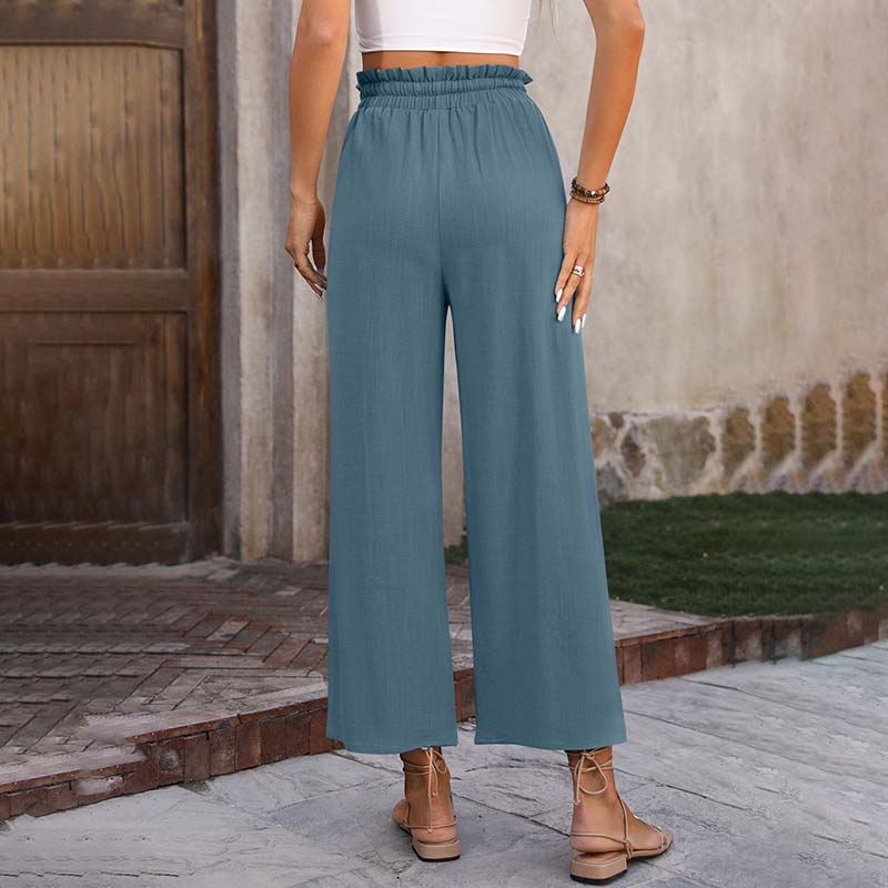 Frau in blauer, hoch taillierter, weiter Hose aus Leinen, steht vor Holztür. Sommermode, lässiger Stil, bequeme Damenbekleidung.