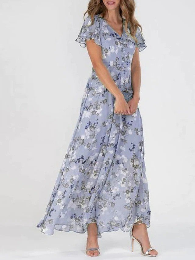 Frau in langem, fließendem, blumigem Kleid in Pastellfarben, steht aufrecht, trägt silberne High Heels. Elegantes Sommeroutfit, modisch, feminin.