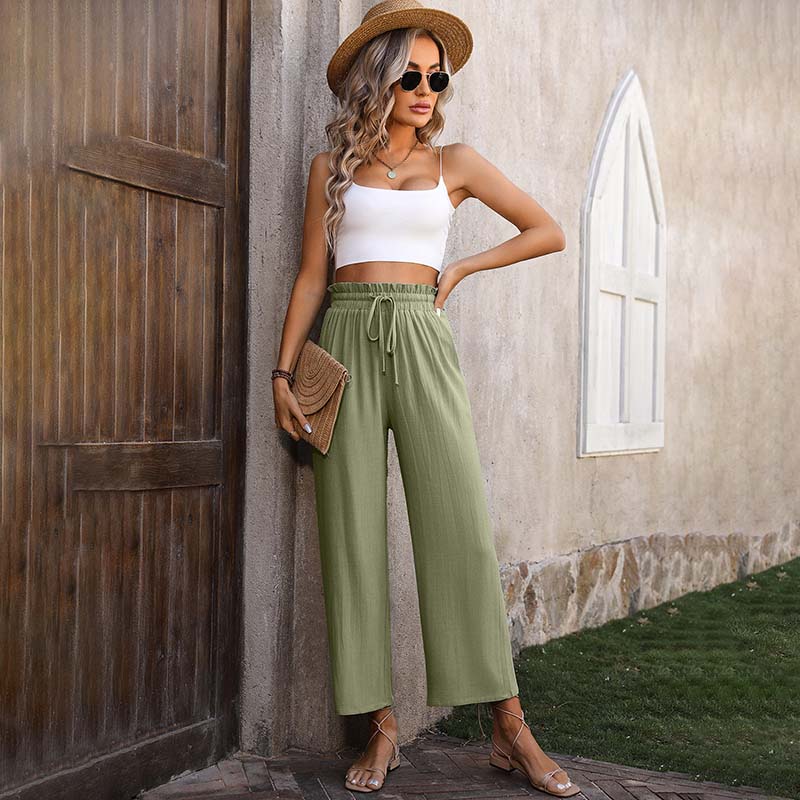 Frau in Sommeroutfit mit Strohhut, weißem Top und grüner Hose lehnt an Wand. Mode, Sommerkleidung, lässiger Stil, Outdoor-Fotografie.