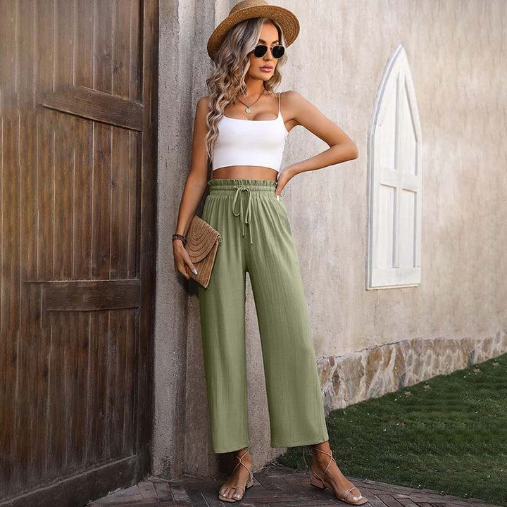 Frau in Sommeroutfit mit Strohhut, weißem Top und grüner Hose lehnt an Wand. Mode, Sommerkleidung, lässiger Stil, Outdoor-Fotografie.