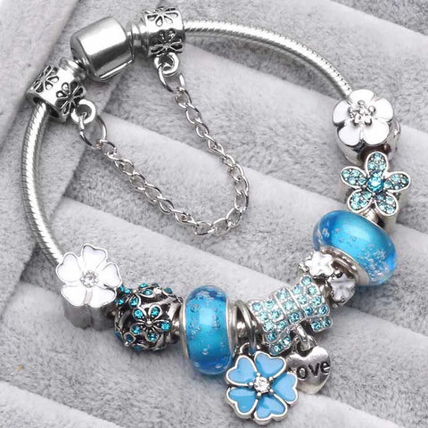 Silbernes Armband mit blauen und silbernen Blumen-Charms, funkelnden Kristallen und Herzanhänger auf grauem Hintergrund. Eleganter Schmuck für Damen.