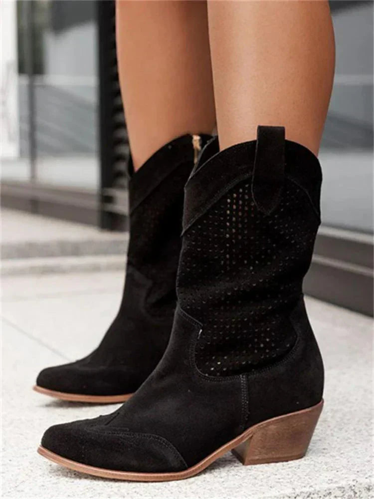 Schwarze Wildleder-Cowboystiefel mit niedrigem Absatz, perforiertem Design und seitlichen Schlaufen, getragen von einer Person auf hellem Boden.