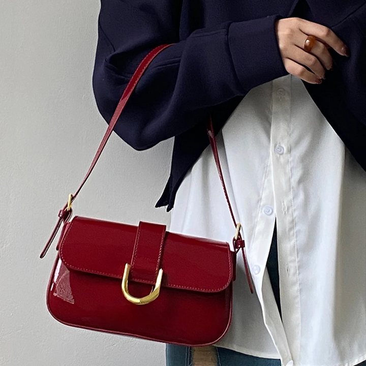 Damenhandtasche aus rotem Lackleder mit goldener Schnalle, getragen von Person in weißer Bluse und dunklem Blazer. Elegante Modeaccessoire.