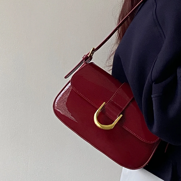 Dunkelrote Umhängetasche aus Leder mit goldener Schnalle, getragen von Person in schwarzem Oberteil. Elegante Damenhandtasche, modisches Accessoire.