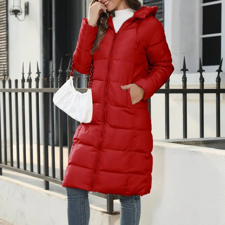 Frau in rotem Daunenmantel, steht vor einem Zaun, trägt weiße Handtasche und Jeans. Wintermode, stilvoll, warm, Outdoor-Bekleidung.