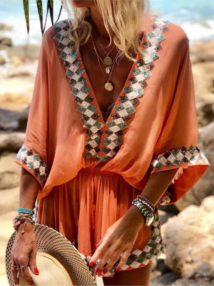 Frau in orangefarbenem Kaftan mit geometrischem Muster am Strand, trägt Schmuck und hält einen Strohhut. Sommermode, Strandkleidung, Boho-Stil.