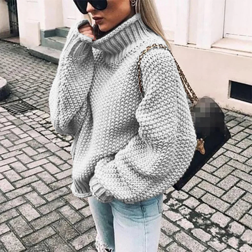 Frau in grauem Strickpullover mit Rollkragen, zerrissene Jeans, Sonnenbrille und schwarzer Tasche auf gepflasterter Straße. Modetrend Herbst/Winter.