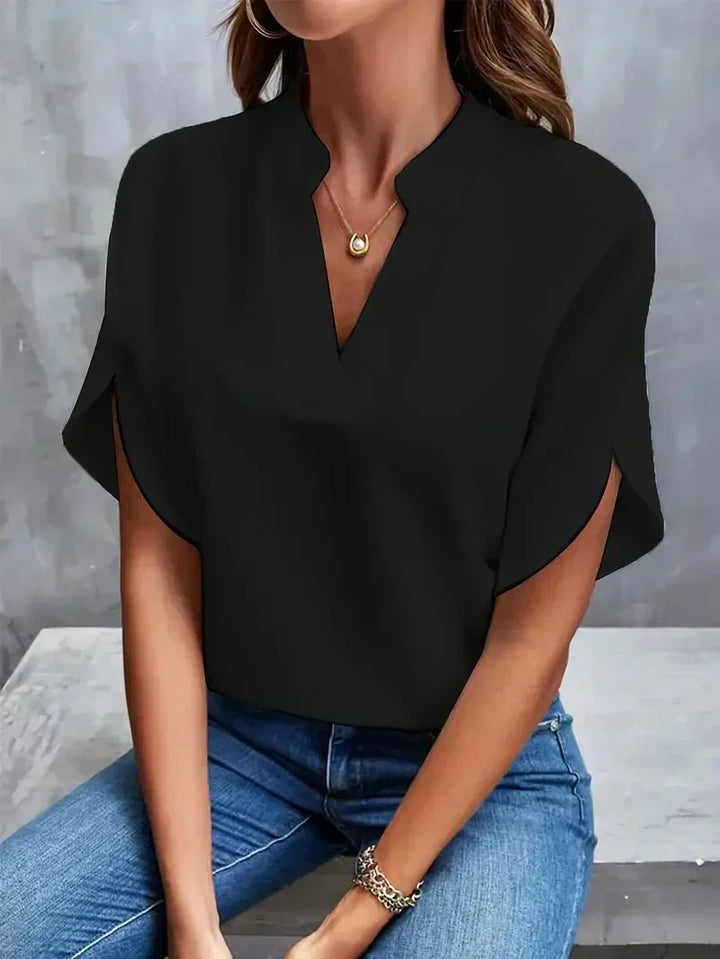 Frau in schwarzer Bluse mit V-Ausschnitt und blauer Jeans, sitzt vor grauem Hintergrund. Modetrend, elegante Damenbekleidung, lässiger Stil.