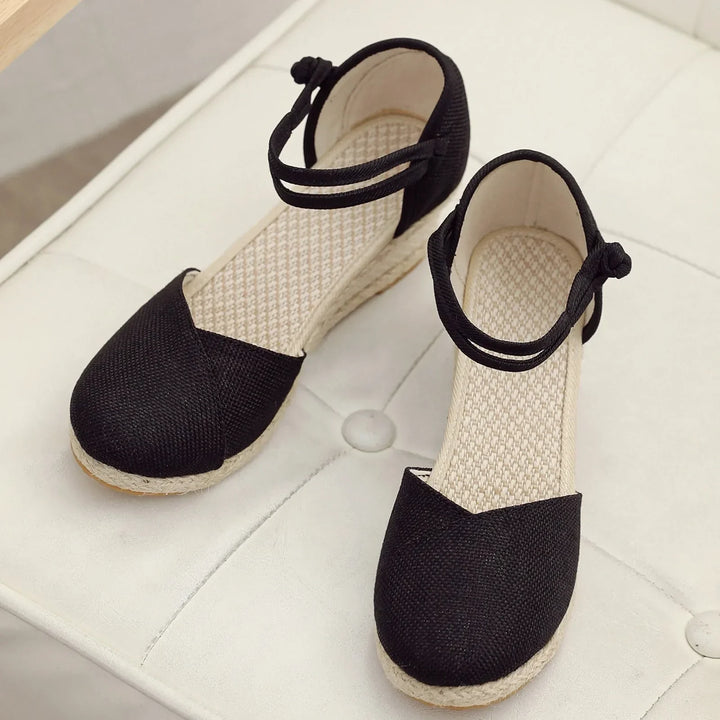 Schwarze Damen-Espadrilles mit Keilabsatz und Knöchelriemen auf weißem Polsterhocker. Modische Sommerschuhe, ideal für Freizeit und Strand.