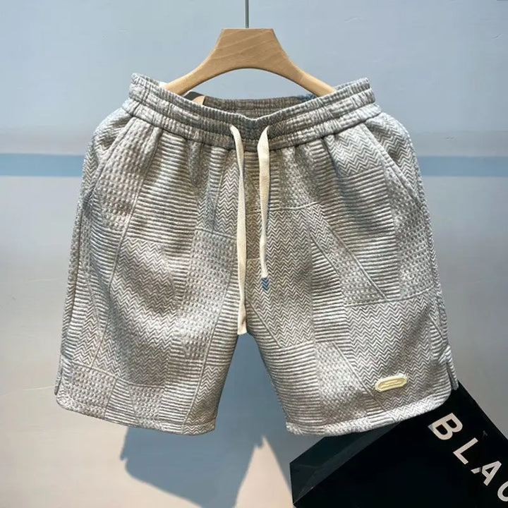 Graue Herren-Shorts mit geometrischem Muster, Kordelzug und elastischem Bund, ausgestellt auf einem Holzkleiderbügel. Ideal für Sommermode.