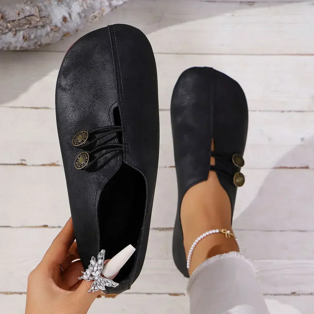 Schwarze, minimalistische Damen-Slipper mit dekorativen Knöpfen, getragen auf weißem Holzboden. Elegante, bequeme Schuhe für den Alltag.