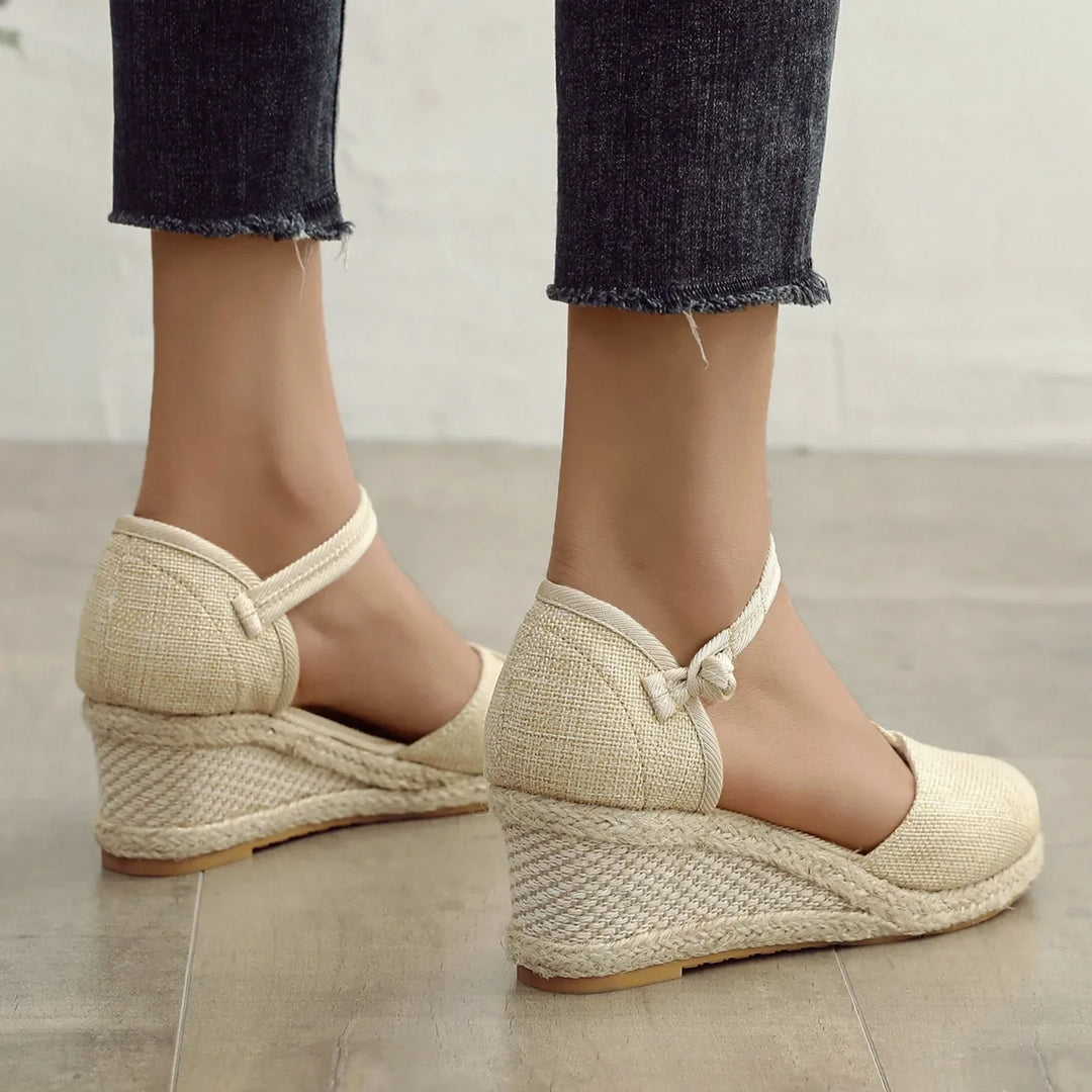 Beige Espadrilles mit Keilabsatz und Knöchelriemen, getragen von einer Person in dunklen Jeans. Modische Sommerschuhe auf Holzboden.
