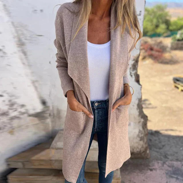 Frau in beiger Strickjacke und weißem Top, kombiniert mit blauen Jeans, steht vor rustikalem Hintergrund. Modetrend, lässiger Herbstlook.