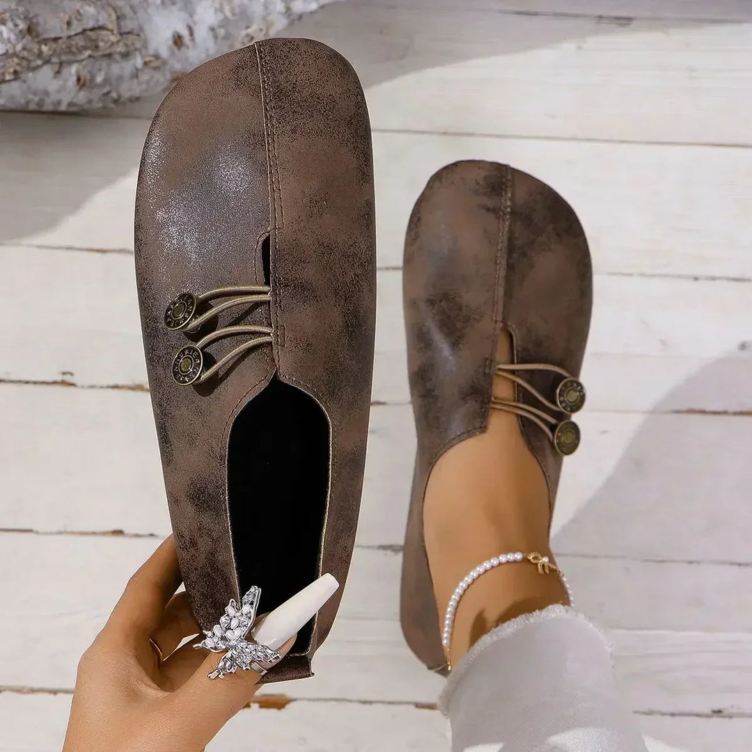 Braune Damen-Loafers mit dekorativen Knöpfen und elastischen Bändern, getragen auf weißem Holzboden. Elegante, bequeme Freizeitschuhe für den Alltag.