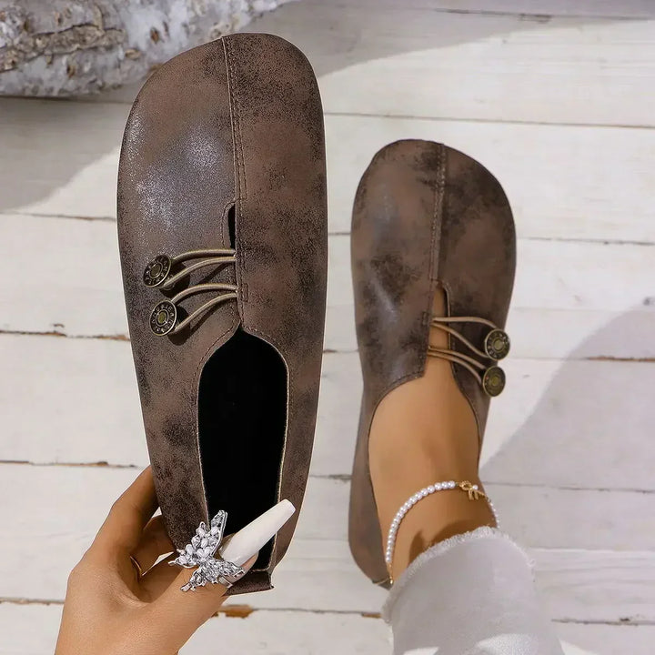 Braune Damen-Loafers mit dekorativen Knöpfen und elastischen Bändern, getragen auf weißem Holzboden. Elegante, bequeme Freizeitschuhe für den Alltag.