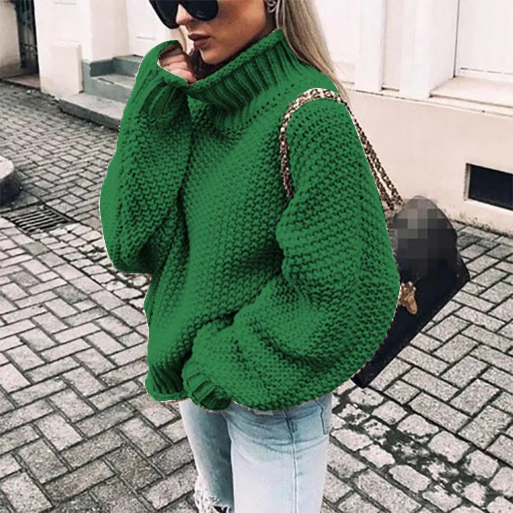 Frau mit grünem Strickpullover und Sonnenbrille, trägt schwarze Tasche, steht auf gepflasterter Straße. Modetrend, Herbstmode, Streetstyle.