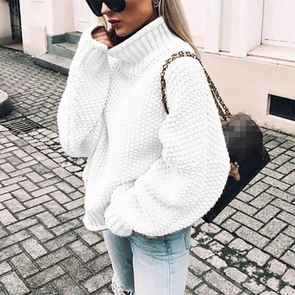 Frau in weißem Strickpullover und Sonnenbrille, trägt schwarze Tasche, steht auf gepflasterter Straße. Modetrend, Wintermode, Streetstyle.