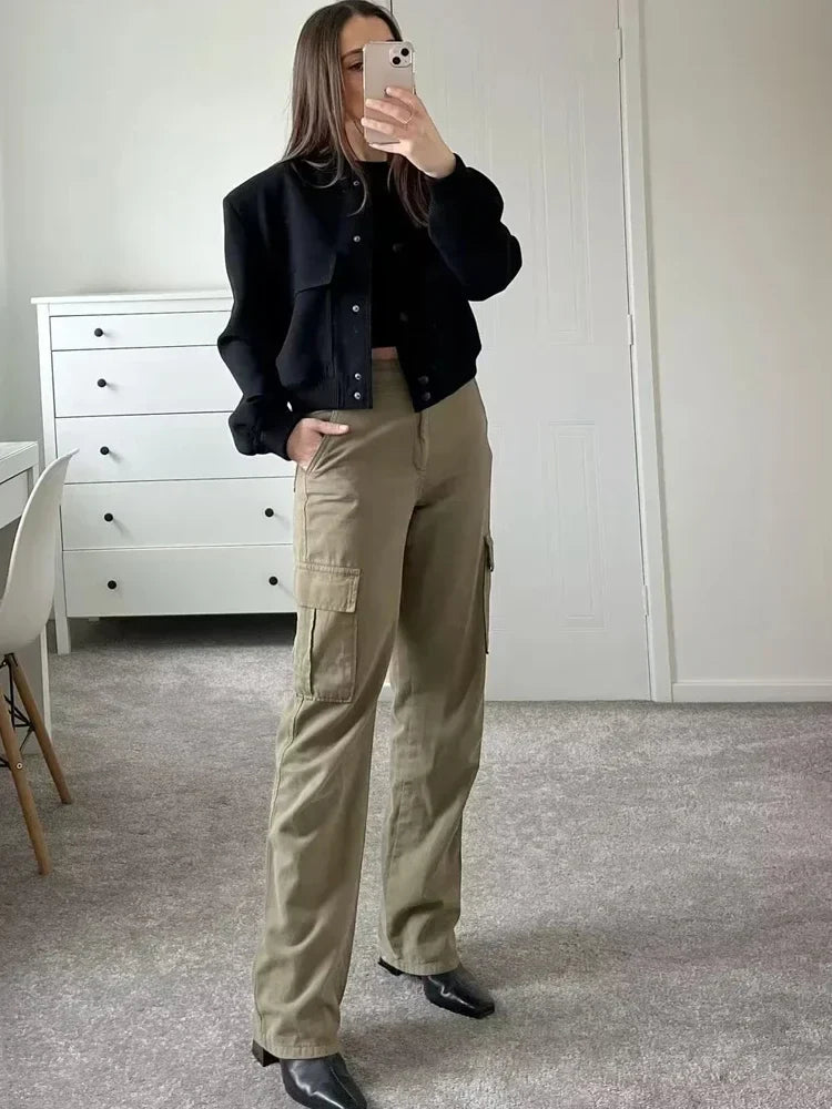 Frau in schwarzer Jacke und khakifarbenen Cargohosen steht in modernem Zimmer, hält Smartphone, modischer Streetstyle-Look, Innenraum.