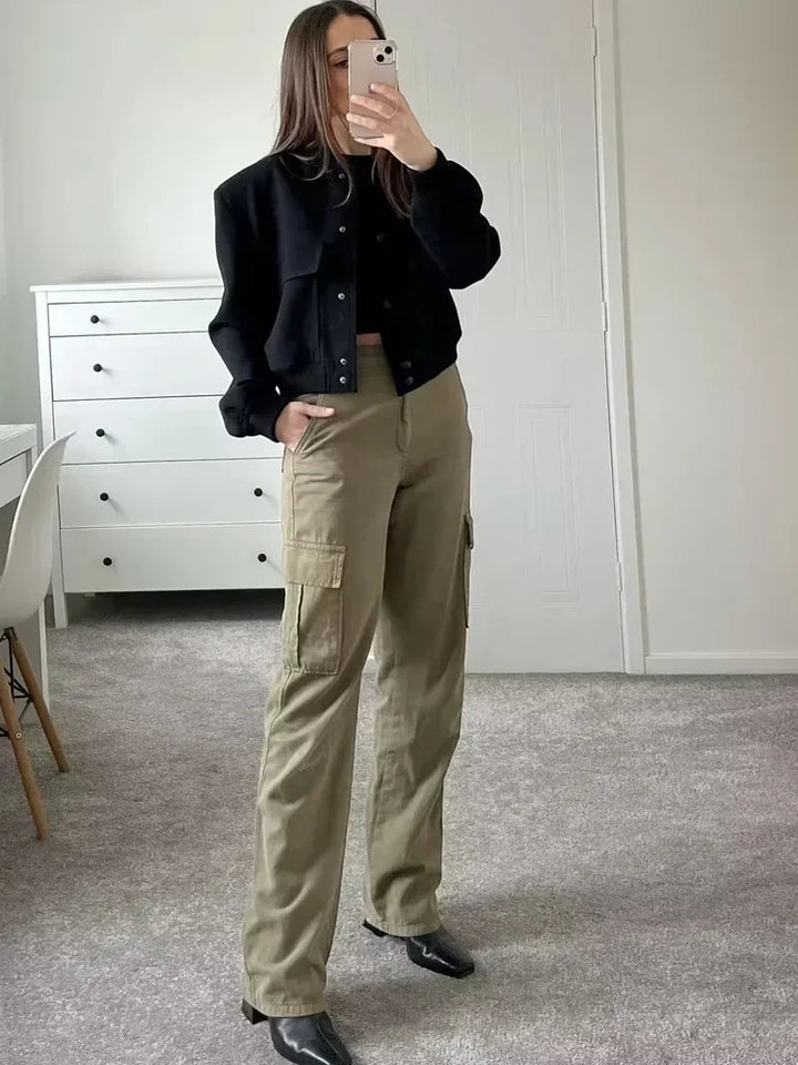 Frau in schwarzer Jacke und khakifarbenen Cargohosen steht in modernem Zimmer, hält Smartphone, modischer Streetstyle-Look, Innenraum.
