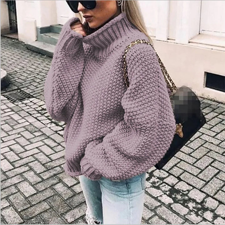 Frau in lila Strickpullover und blauer Jeans, trägt schwarze Sonnenbrille und Handtasche, steht auf gepflasterter Straße.