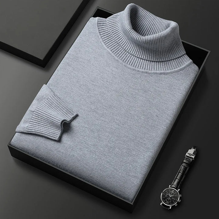 Grauer Rollkragenpullover aus Kaschmir in schwarzer Geschenkbox, elegant gefaltet, neben einer stilvollen Armbanduhr auf schwarzem Hintergrund.