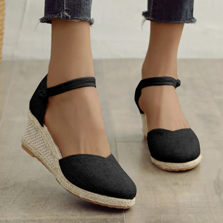 Schwarze Espadrilles mit Keilabsatz und Knöchelriemen, getragen von einer Person auf Holzboden. Modische Sommerschuhe für Damen.