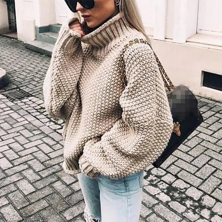 Frau in beigem Strickpullover und blauer Jeans auf Kopfsteinpflasterstraße, trägt schwarze Sonnenbrille und braune Handtasche.