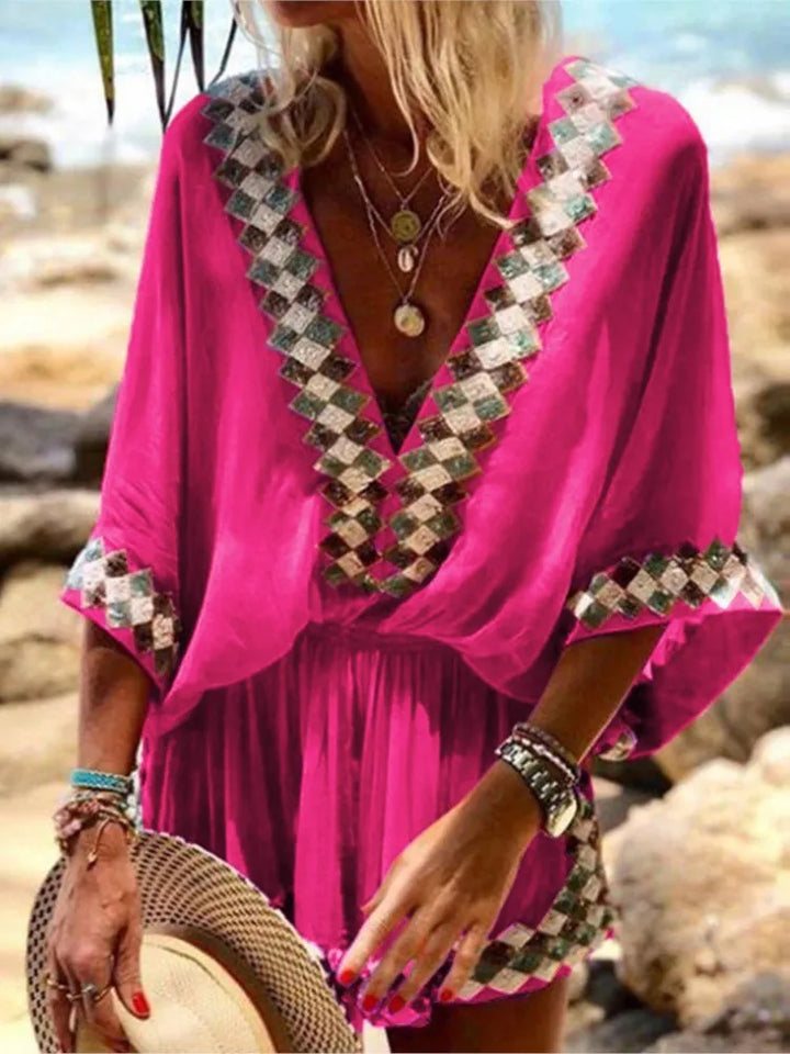Frau in pinkem Kaftan mit V-Ausschnitt und geometrischem Muster, hält Strohhut, trägt Armbänder und Ketten, am Strand. Sommermode, Strandkleidung.