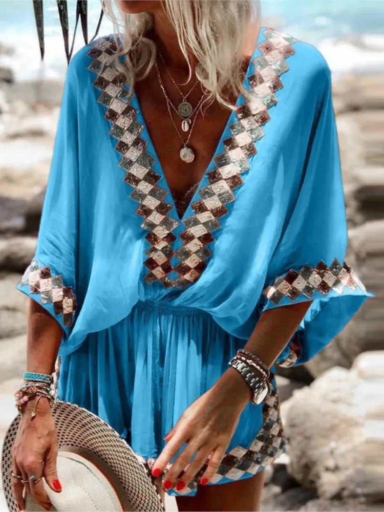 Frau in blauem Strandkleid mit geometrischem Muster, hält Strohhut. Sommermode, Strandoutfit, Boho-Stil, Schmuck, Meereshintergrund.