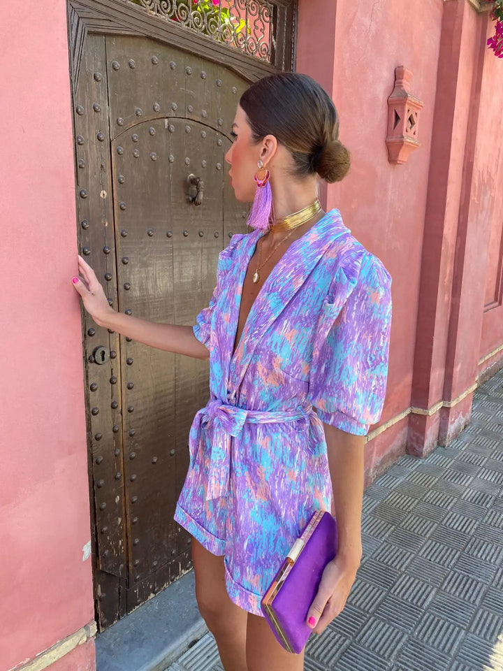 Frau in lila-pinkem Kleid mit goldenen Accessoires vor antiker Holztür, hält lila Clutch. Elegante Mode, Streetstyle, Sommeroutfit.