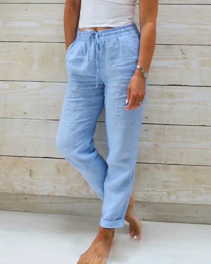 Frau in hellblauer Leinenhose und weißem Top vor Holzwand. Bequeme Sommermode, lässiger Stil, Damenbekleidung, Freizeitlook.