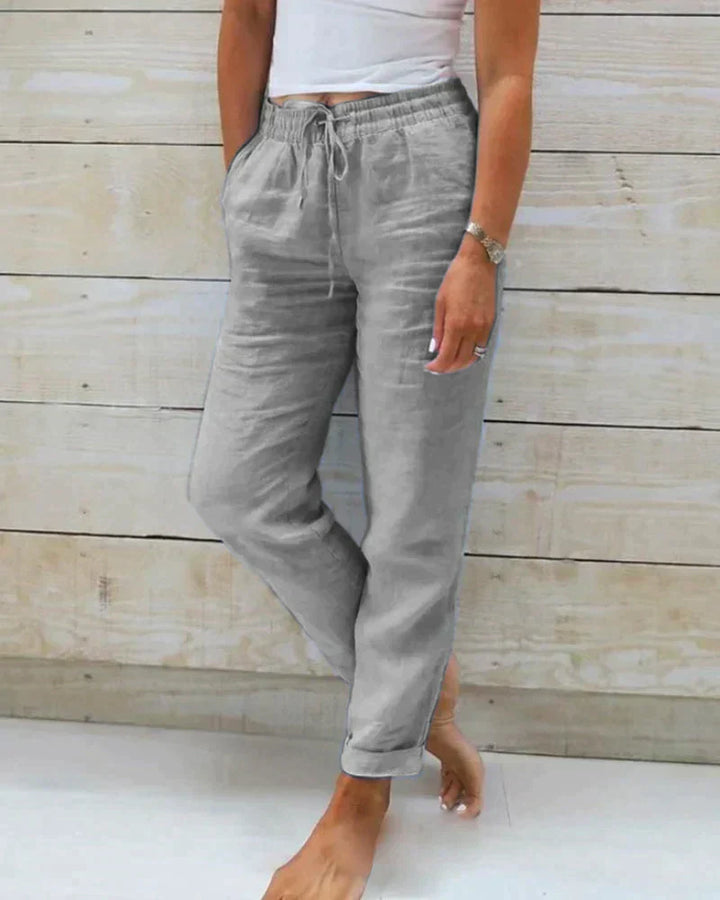 Frau in grauer Leinenhose und weißem Top vor Holzwand, lässiger Sommerlook, Mode für Damen, bequeme Freizeitkleidung, barfuß.