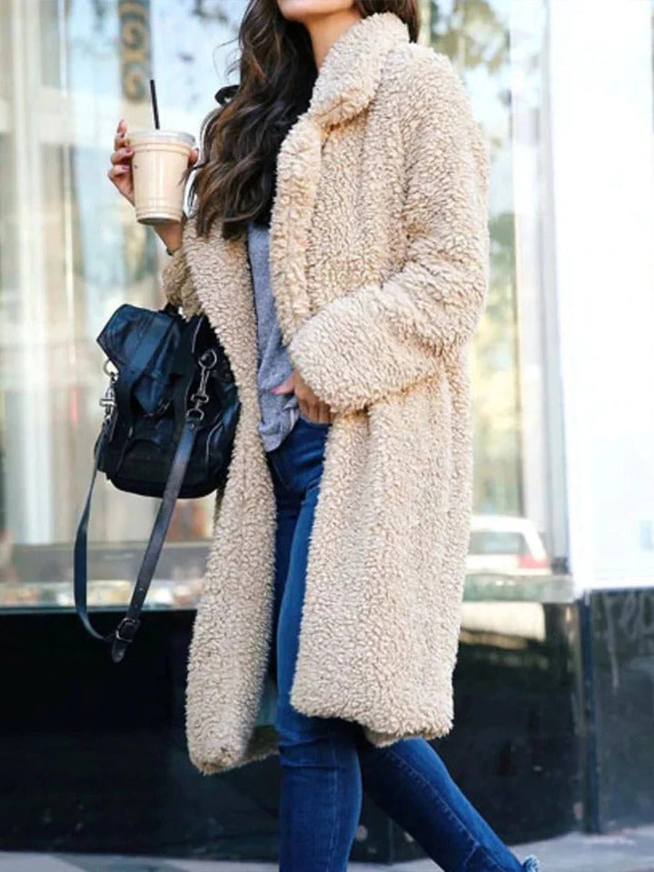 Frau in beigem Teddy-Mantel, hält Eiskaffee und schwarze Tasche, trägt blaue Jeans. Modetrend Herbst/Winter, urbaner Stil.