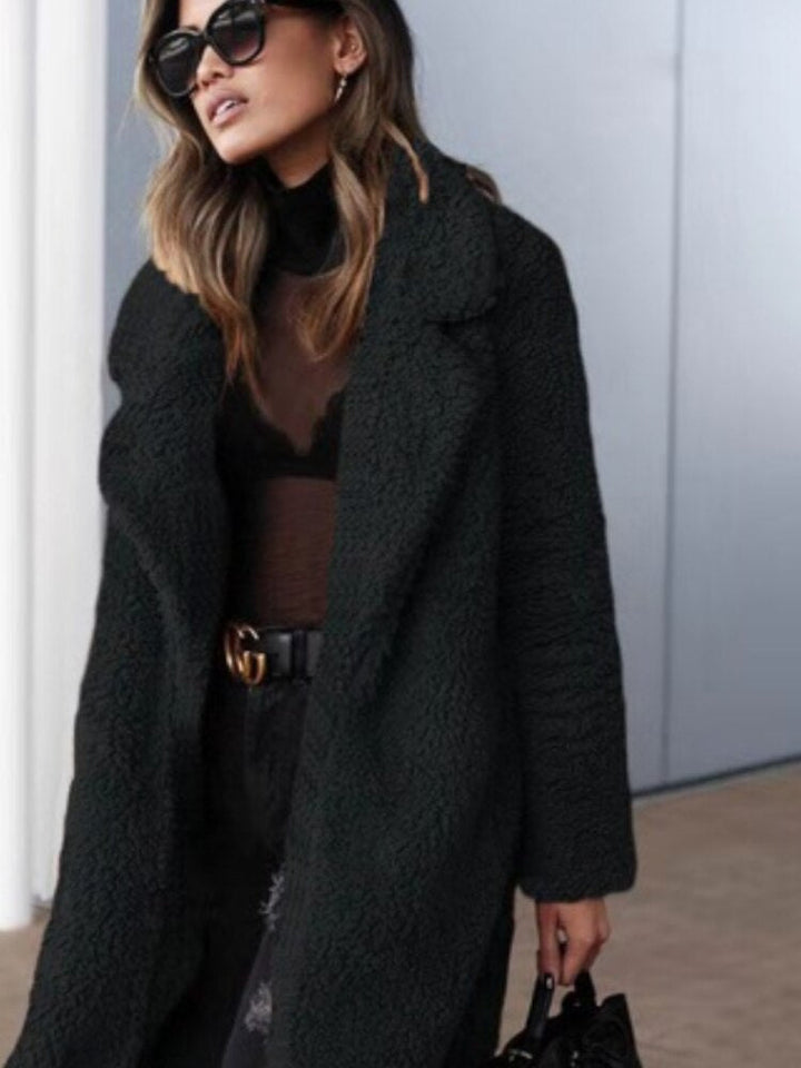 Frau in schwarzem Teddy-Mantel, Sonnenbrille und durchsichtigem Oberteil, modisch elegant gekleidet, hält eine Handtasche. Trendige Herbstmode.