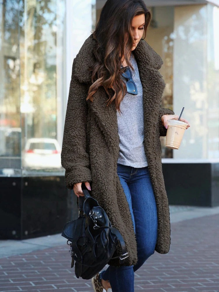 Frau in braunem Teddy-Mantel, grauem Pullover und blauer Jeans, hält Eiskaffee und schwarze Handtasche. Straßenmode, Herbstlook, lässiger Stil.