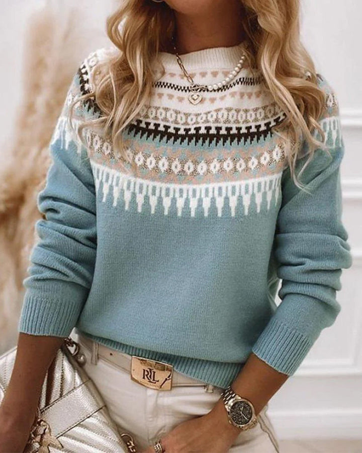 Frau in blauem Norwegerpullover mit weißem Muster, trägt goldene Uhr und Kette, hält silberne Tasche. Modetrend Herbst/Winter 2023.