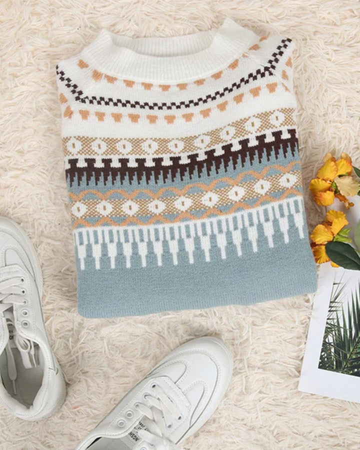 Gefalteter Strickpullover mit nordischem Muster in Weiß, Braun und Blau auf beigem Teppich, neben weißen Sneakern und gelben Blumen.