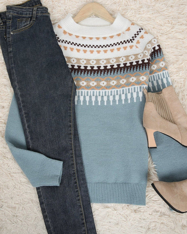 Blauer Strickpullover mit Norwegermuster, kombiniert mit dunkelblauer Jeans und beigen Stiefeletten auf einem cremefarbenen Teppich.